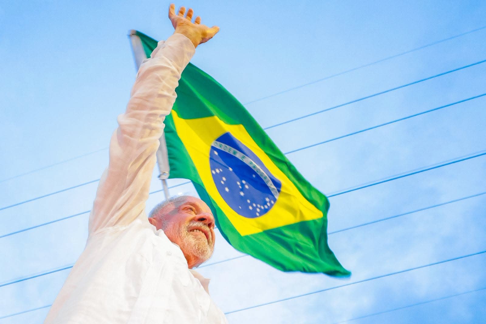 Lula, de camisa branca, com o braço direito erguido diante da bandeira do Brasil